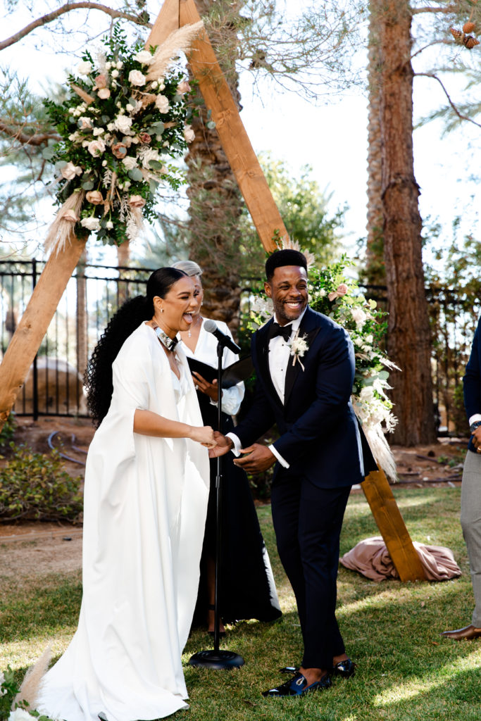 Bride and groom laughing during their wedding ceremony with triangle, wooden arbor and neutral floral arrangement in the background.