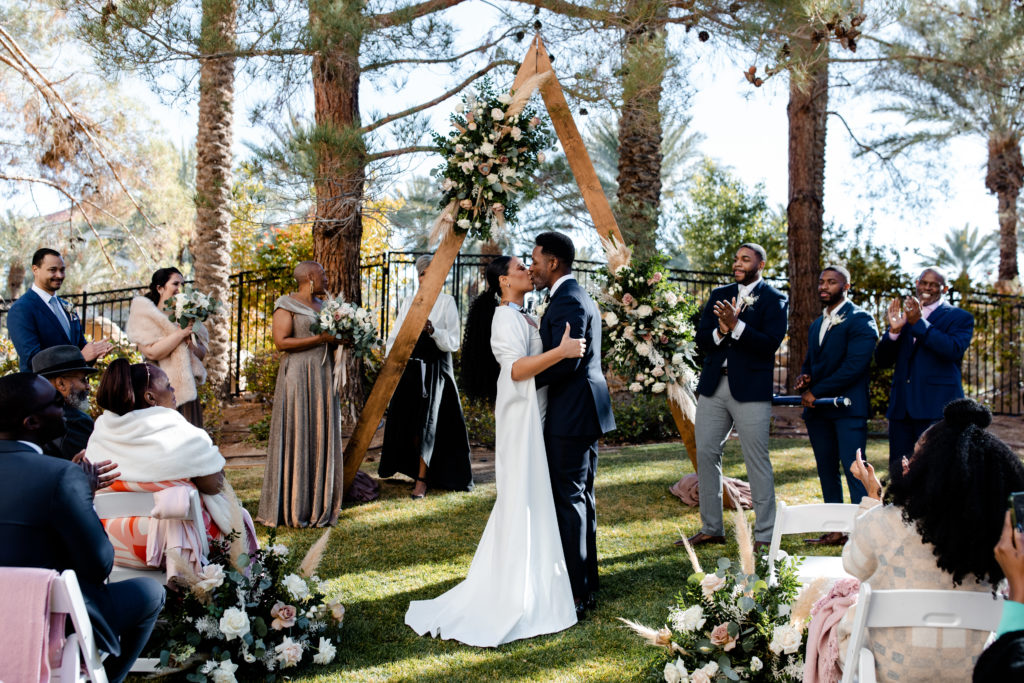 First kiss at this JW Marriott Las Vegas Wedding in front of a triangle arbor with neutral florals.
