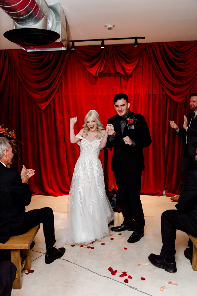 The bride and groom walk back down the aisle hand in hand, grinning with joy as their guests cheer them on amidst the punk rock memorabilia.