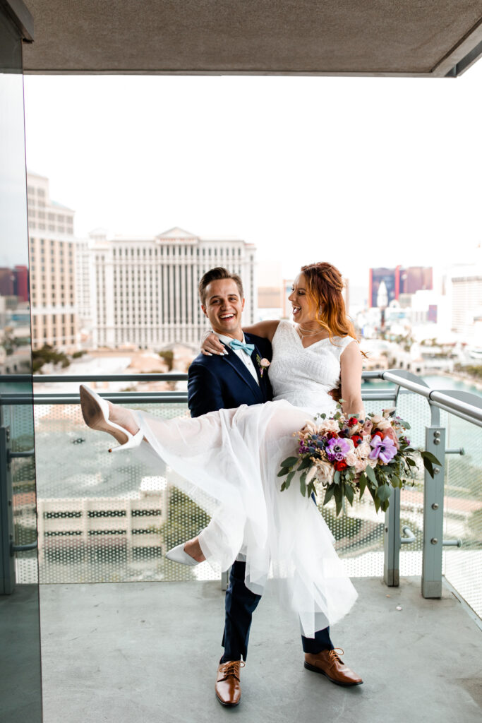 Las Vegas Photographer captures The groom playfully lifts the bride on the balcony of their Cosmopolitan suite, both of them laughing with joy against the backdrop of the Las Vegas skyline.