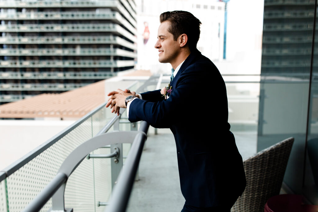 The groom stands confidently on the balcony of the Cosmopolitan suite, with the city's bright lights and energy behind him.