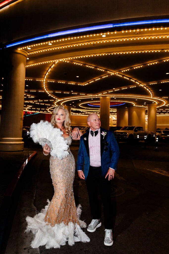 Bride and groom pose under the bright marquee lights outside the Flamingo in Las Vegas, her rhinestone gown and feather fan catching the glow