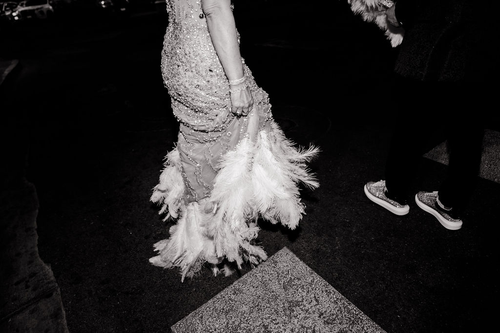 Black and white close-up of the bride’s beaded gown and feathered hem as she walks beside the groom.
