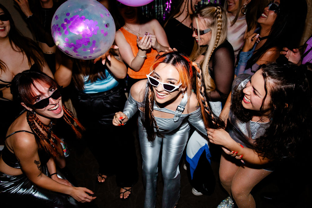 Bride in a silver two-piece set with white sunglasses, surrounded by friends in bold black and metallic outfits