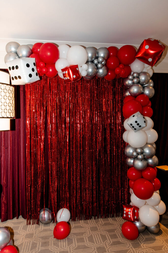 Red, white, and silver balloon arch with dice balloons and metallic fringe backdrop at a Lucky in Love bachelorette party in Las Vegas