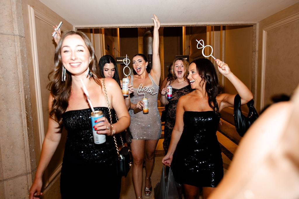 Bride and bridesmaids walking through hotel hallway, holding drinks and laughing, heading out for their Lucky in Love bachelorette party in Las Vegas