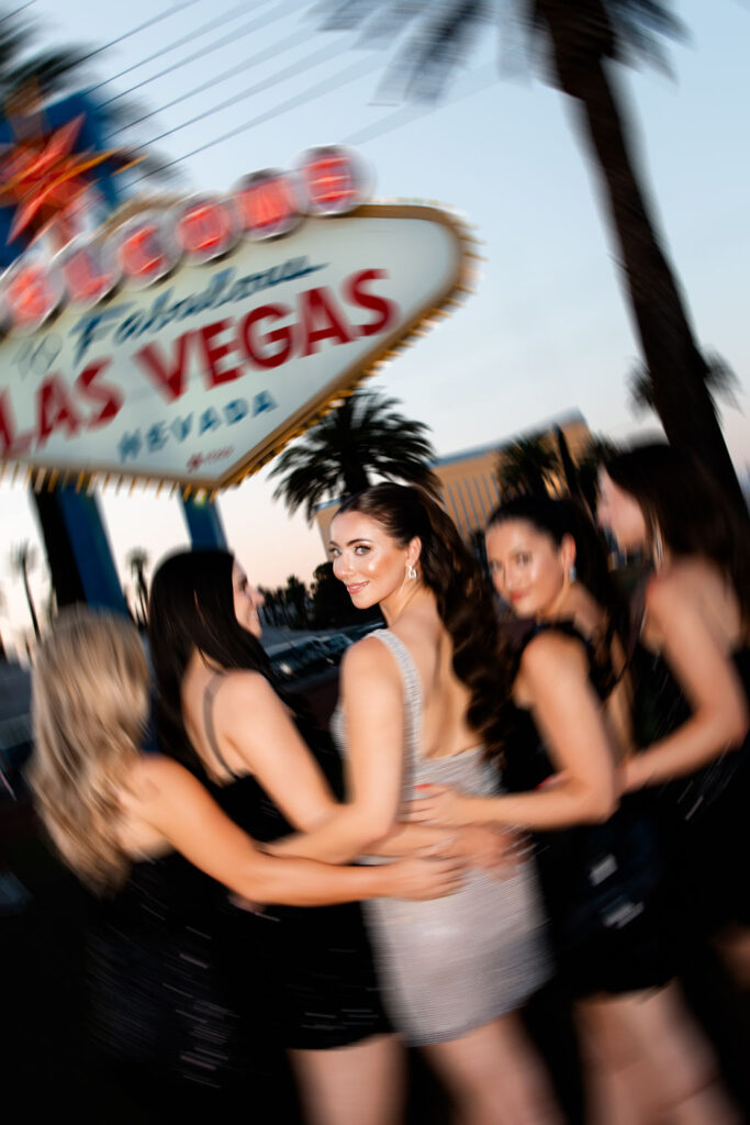 Bride in sparkly silver dress looking back with a smile while standing with friends in front of the Welcome to Fabulous Las Vegas sign