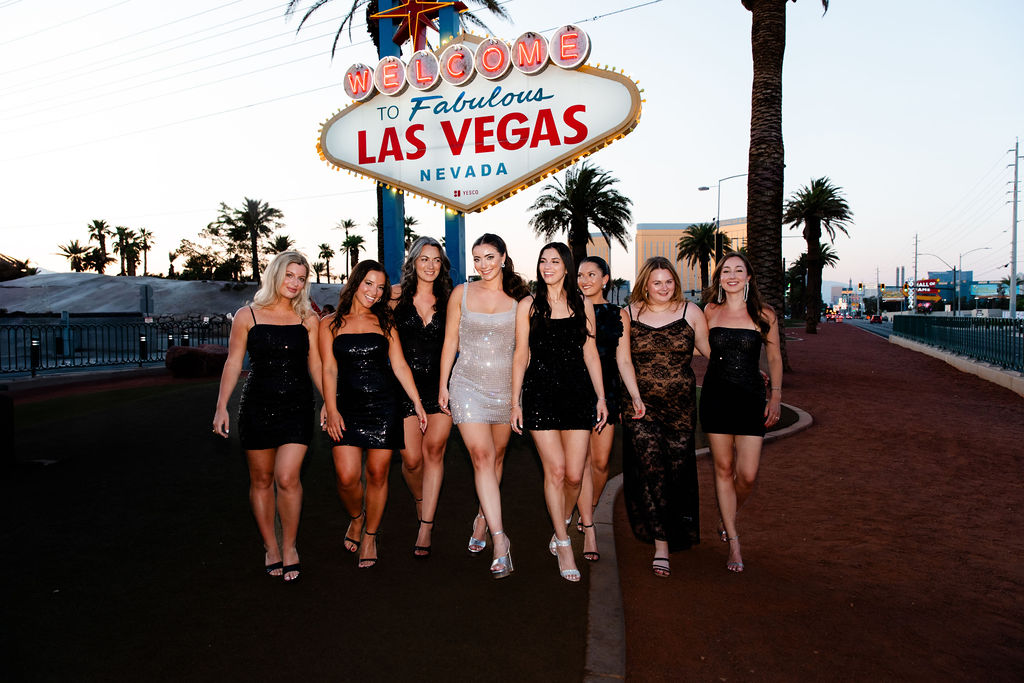 Bride and bridesmaids in black and sparkly dresses walking together at the Welcome to Fabulous Las Vegas sign during a bachelorette weekend.