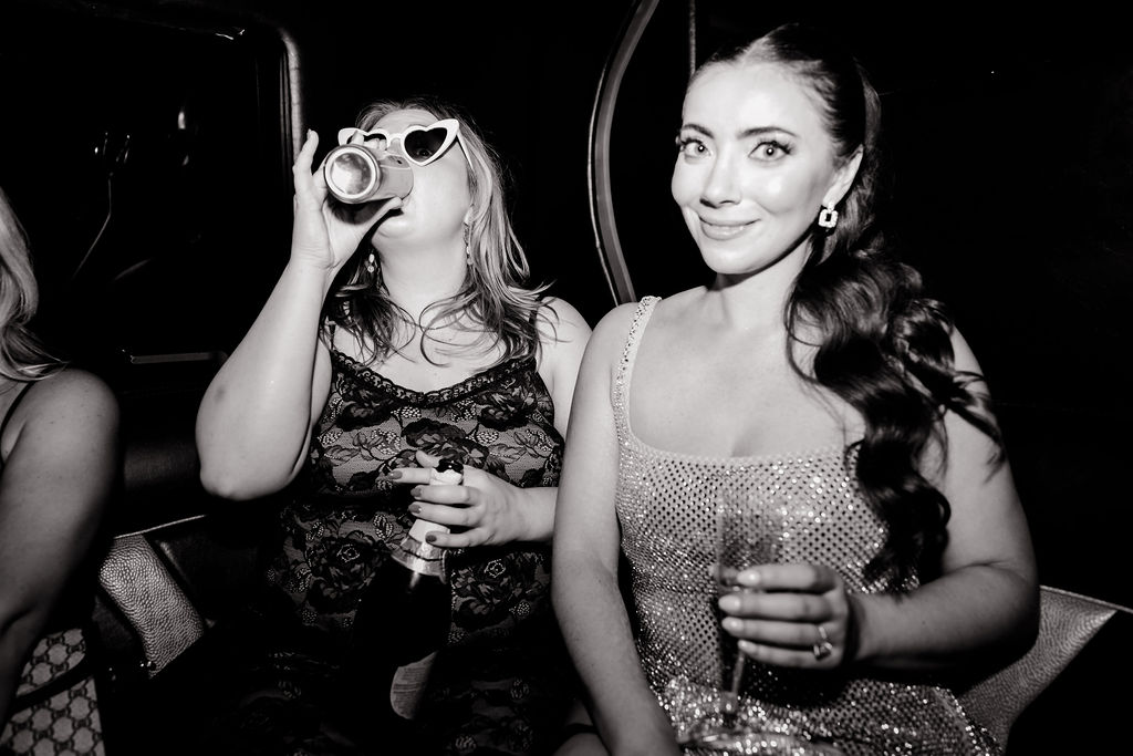 Black and white photo of bride smiling in sparkly dress while friend sips a drink wearing heart sunglasses inside the limo