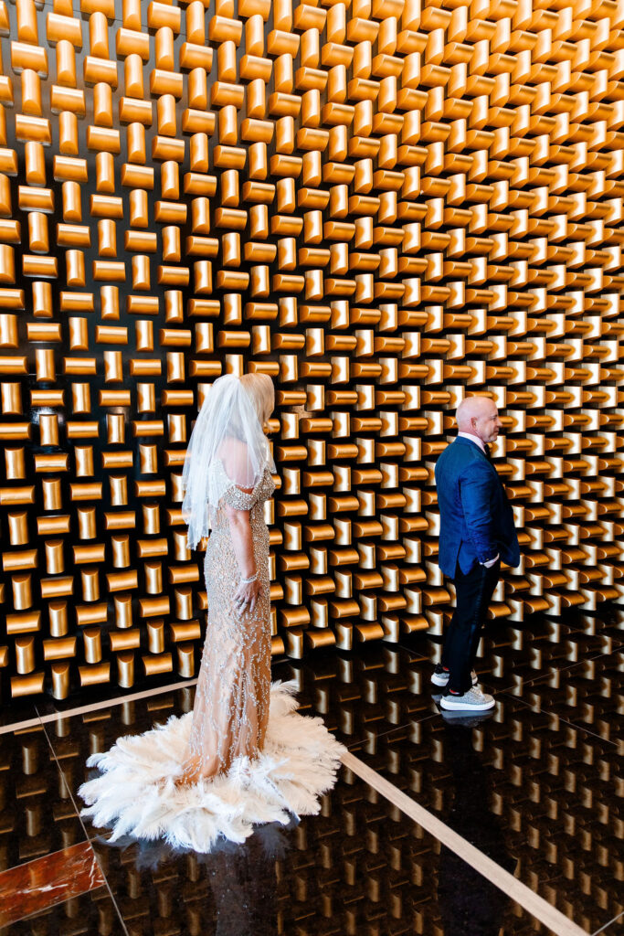 A bride approaches the groom during their first look, standing against the modern gold art wall inside Waldorf Astoria