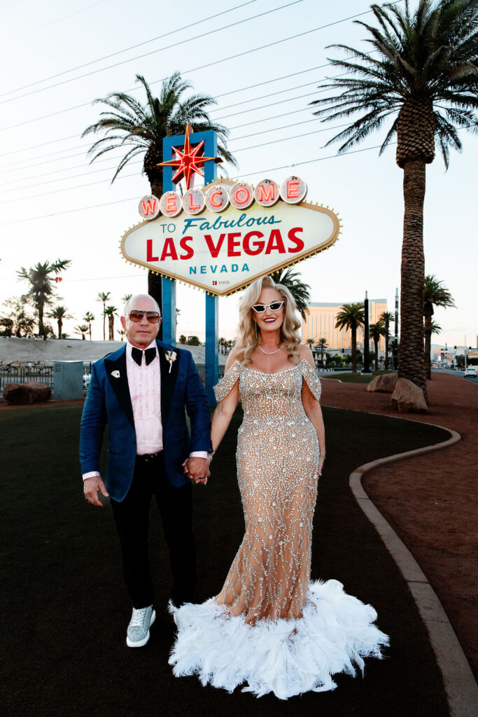 A bride in a glittering gown and groom in a blue suit walk hand in hand beneath the famous neon sign.