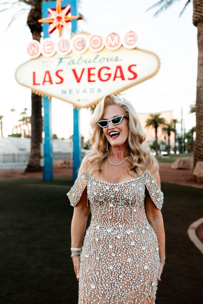 A glamorous bride in sunglasses smiles under the neon sign during her Vegas wedding day portraits.