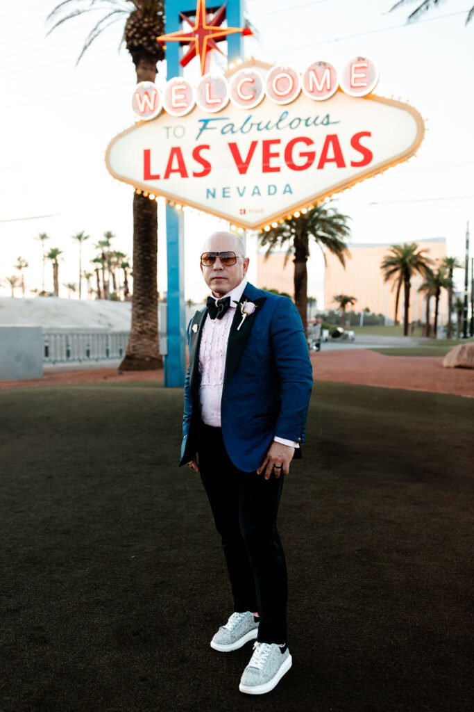 A groom in a patterned blue jacket, pink shirt, and sneakers poses confidently in front of the Welcome to Las Vegas sign