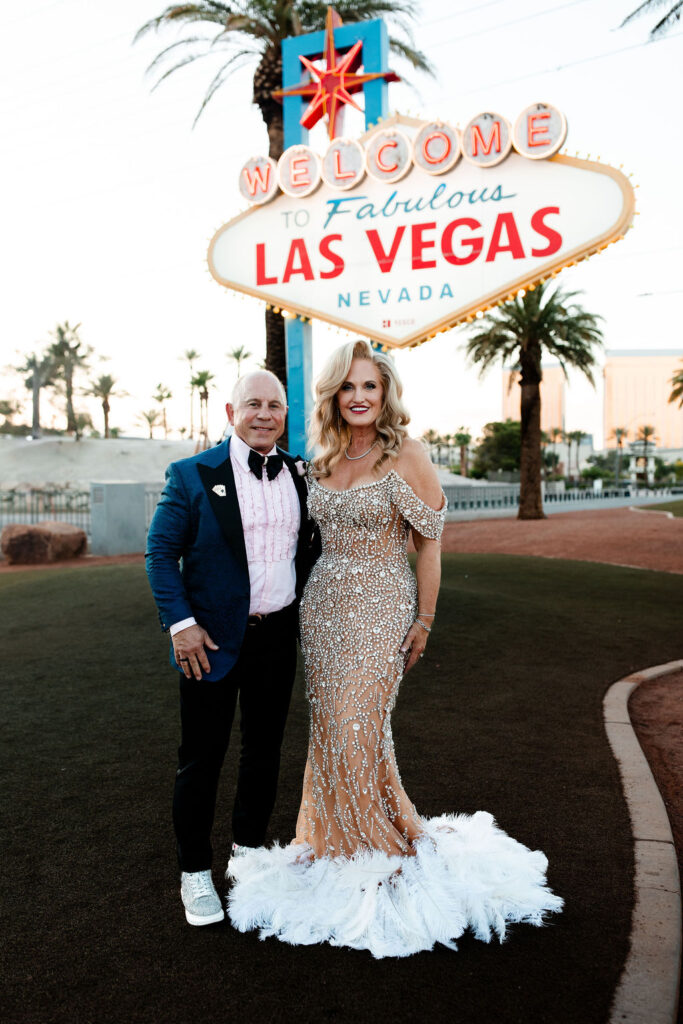 A bride in a sparkling crystal gown with feather details poses with the groom in a blue jacket beneath the Vegas sign