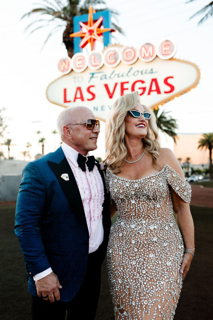 The couple smiles in sunglasses, dressed in glamorous attire, with the iconic Welcome to Las Vegas sign behind them