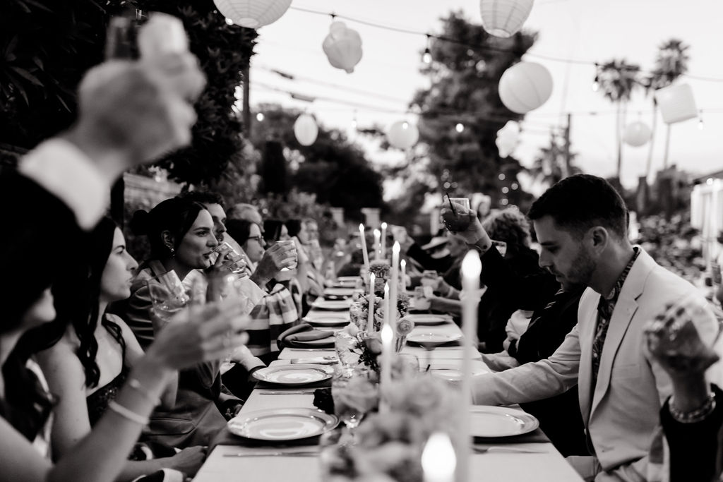 Black and white photo of reception guests raising glasses in a toast at long tables lined with candles.